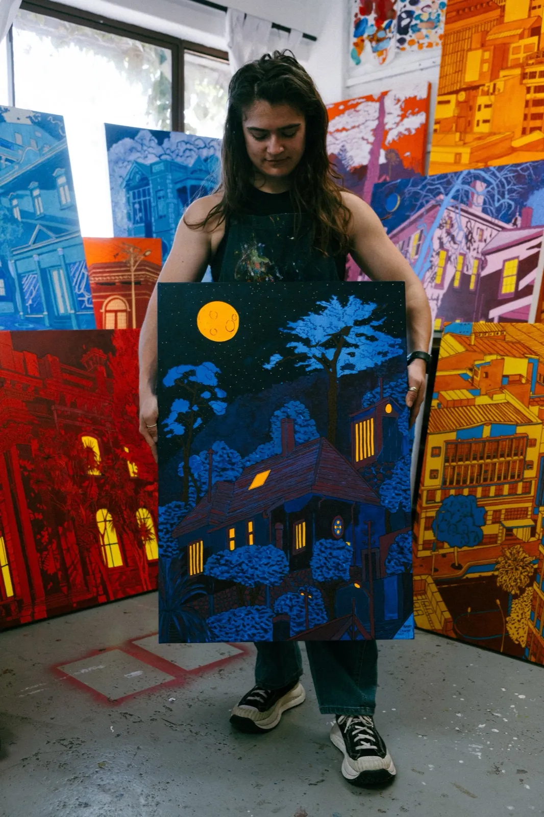 Aimee in her studio holding a night-time painting of a house under a full moon, surrounded by her Cape Town building paintings